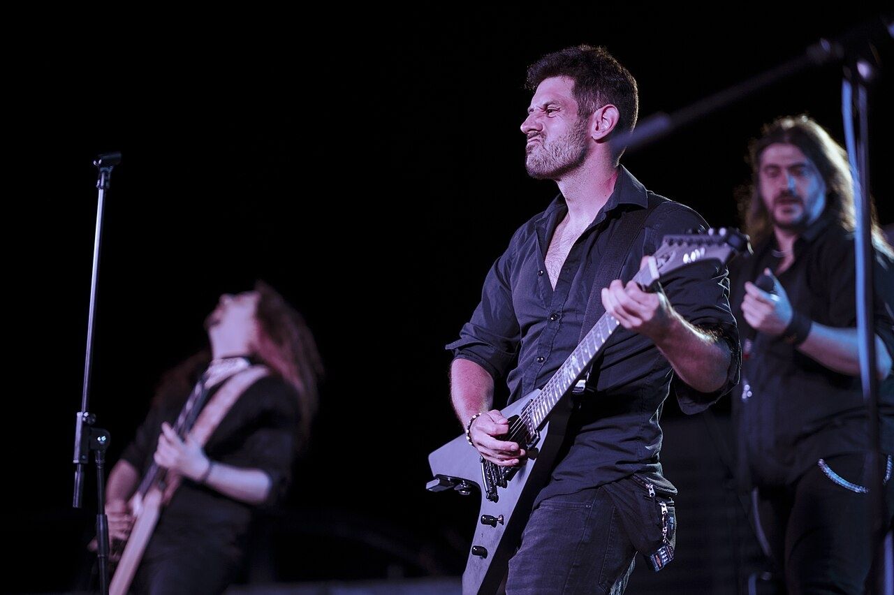 Three musicians play electric guitars during a live outdoor concert. The crowd e